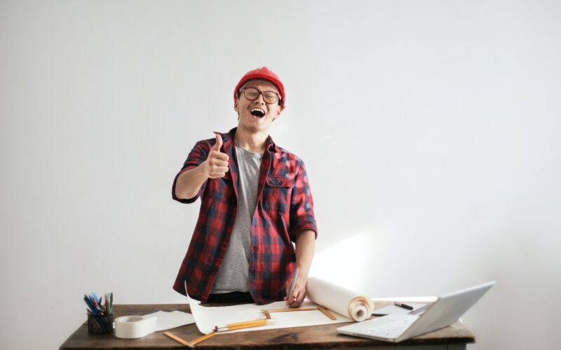 man in hard hat standing over a desk with paperwork giving a thumbs up.